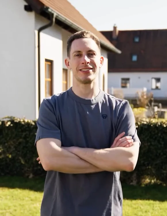 Mann steht mit verschrenkten Armen auf einem Rasen und lächelt in die Kamera.